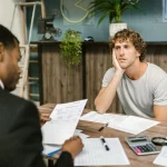 Man in office, appearing stressed during paperwork discussion, sitting opposite professional.