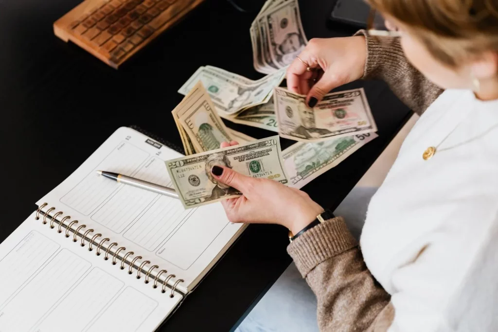 Close-up of hands counting dollar bills with calculator, notebook, and laptop on wooden table.